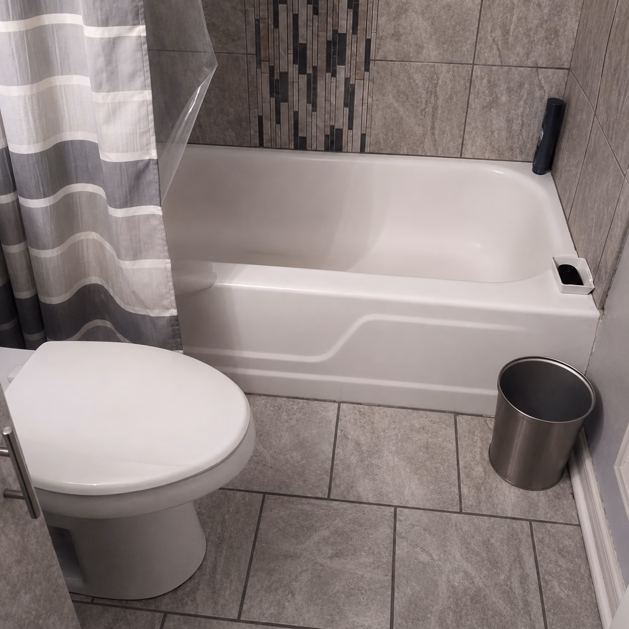 Bathroom with white toilet, bathtub, gray tiled floor and wall, striped shower curtain, and silver trash can.