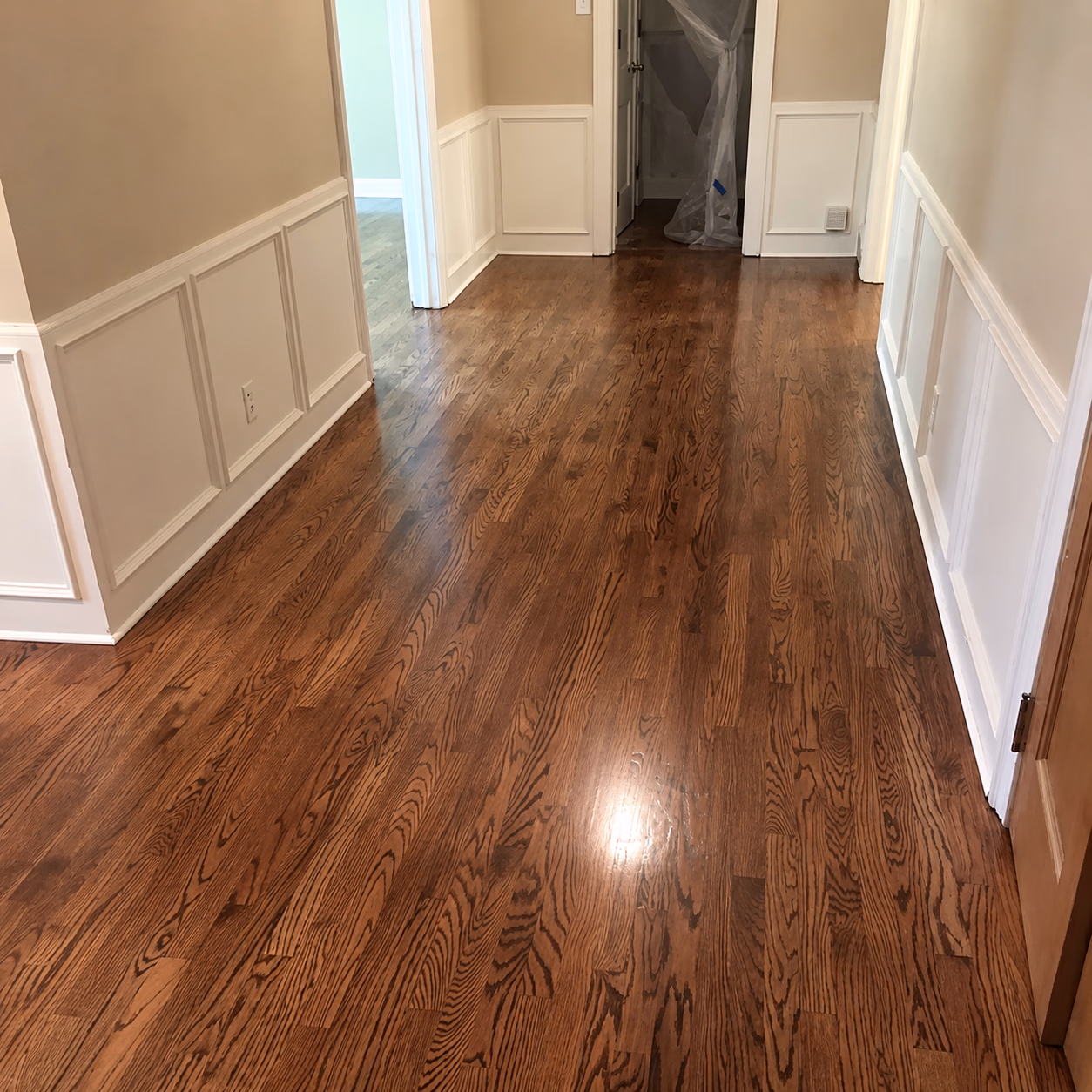 Empty hallway with polished wooden floors, white wainscoting, and beige walls with doors ahead.