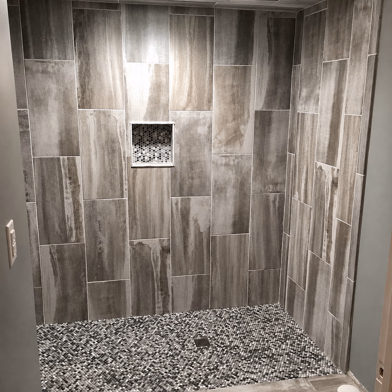 Walk-in shower with large gray tiles on walls and small pebble-patterned tiles on floor.