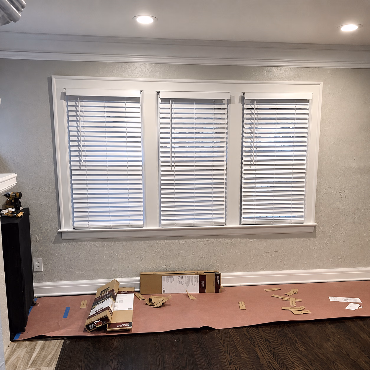 Three closed white blinds on a wall with boxes and scattered packaging on the floor covered by pink protective paper.