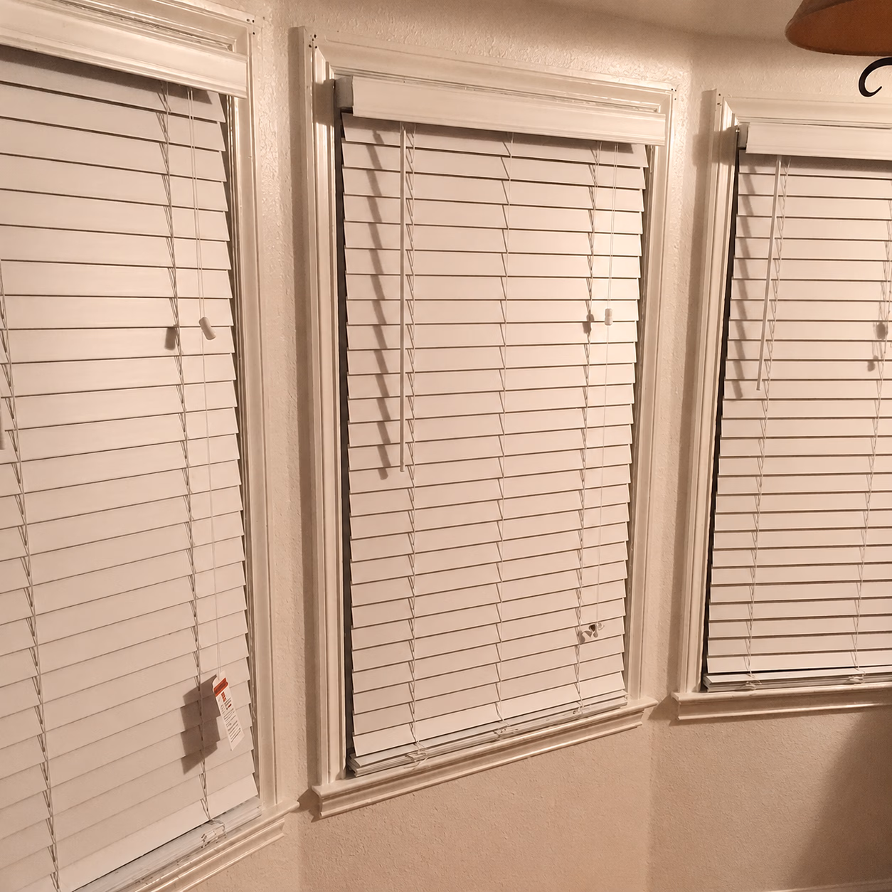 Three white window blinds closed over windows with white trim on beige walls.