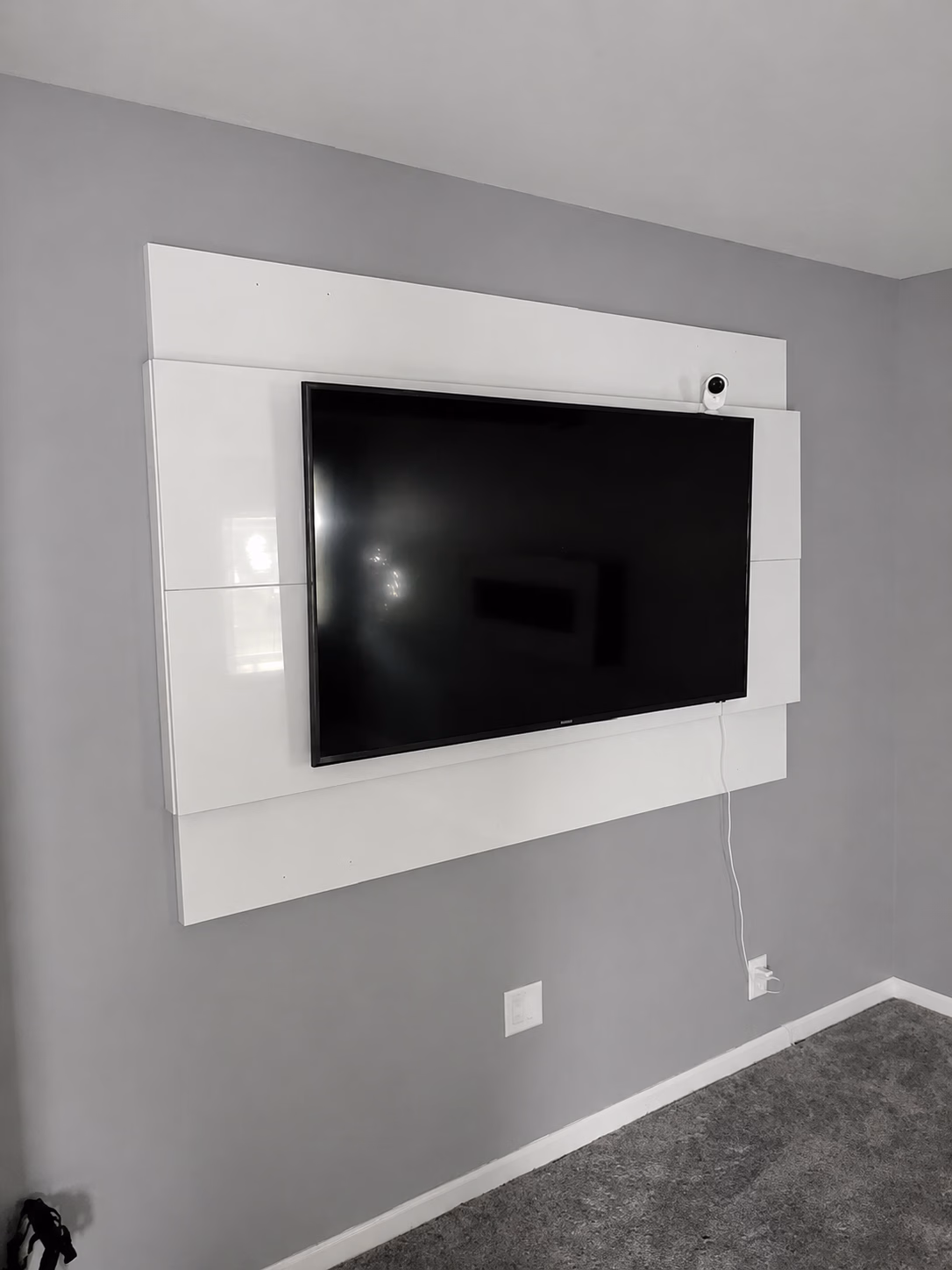Flat screen TV mounted on a white panel against a gray wall with a small security camera above it.