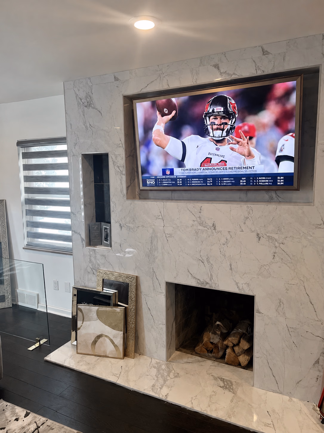 Modern marble fireplace with stacked wood and TV showing Tom Brady announcing retirement.