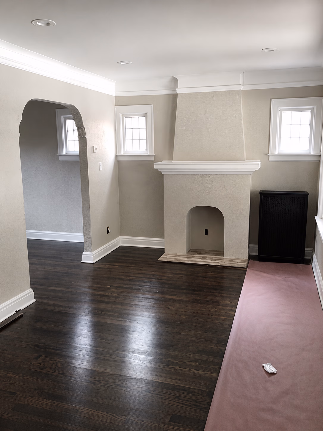 Empty room with dark wood floor, arched doorway, small windows, white fireplace, and pink floor covering.