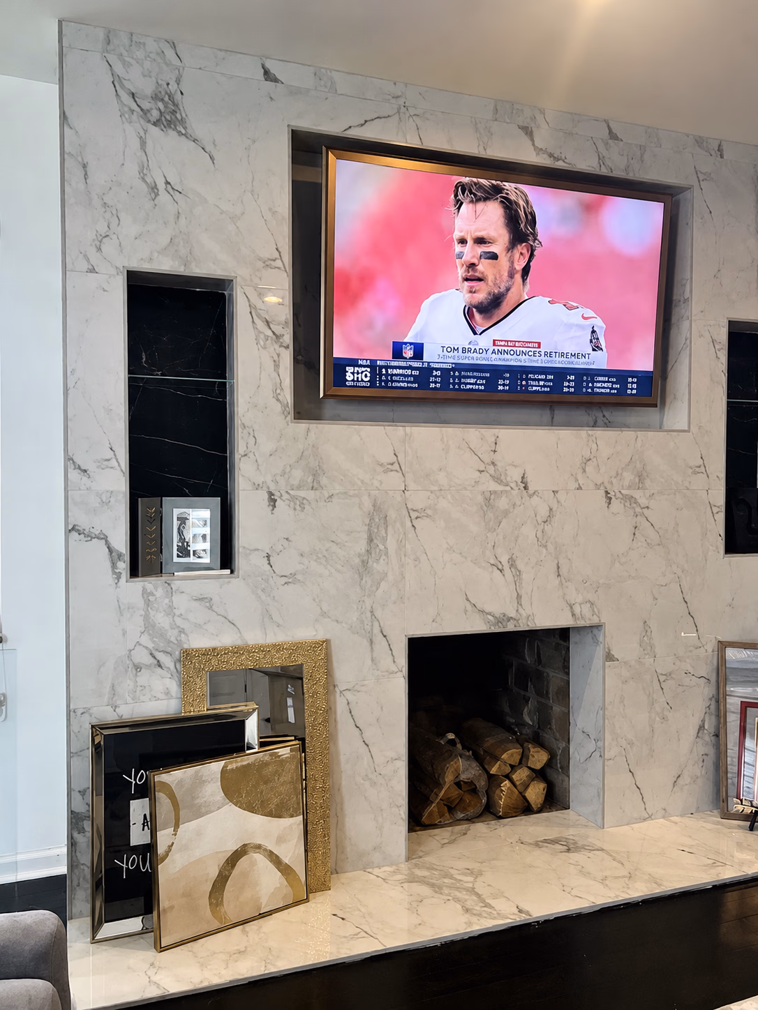 Marble fireplace with stacked firewood and TV above showing Tom Brady announcing retirement.