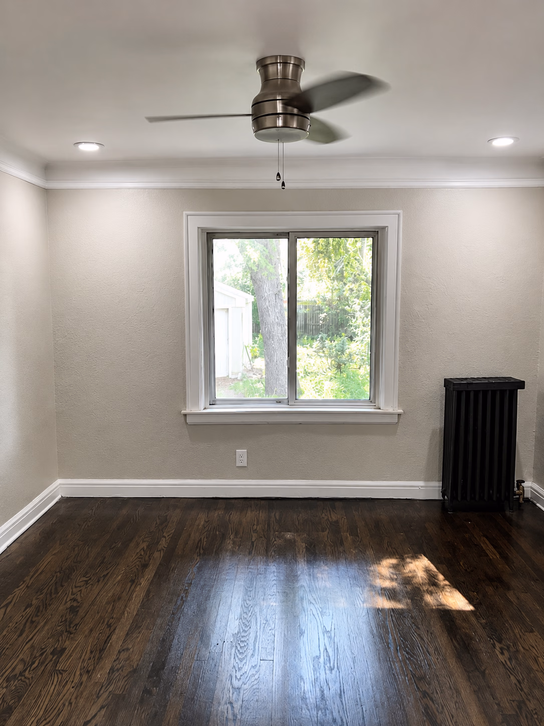 Empty room with dark hardwood floor, beige walls, ceiling fan, window showing backyard, and black radiator.