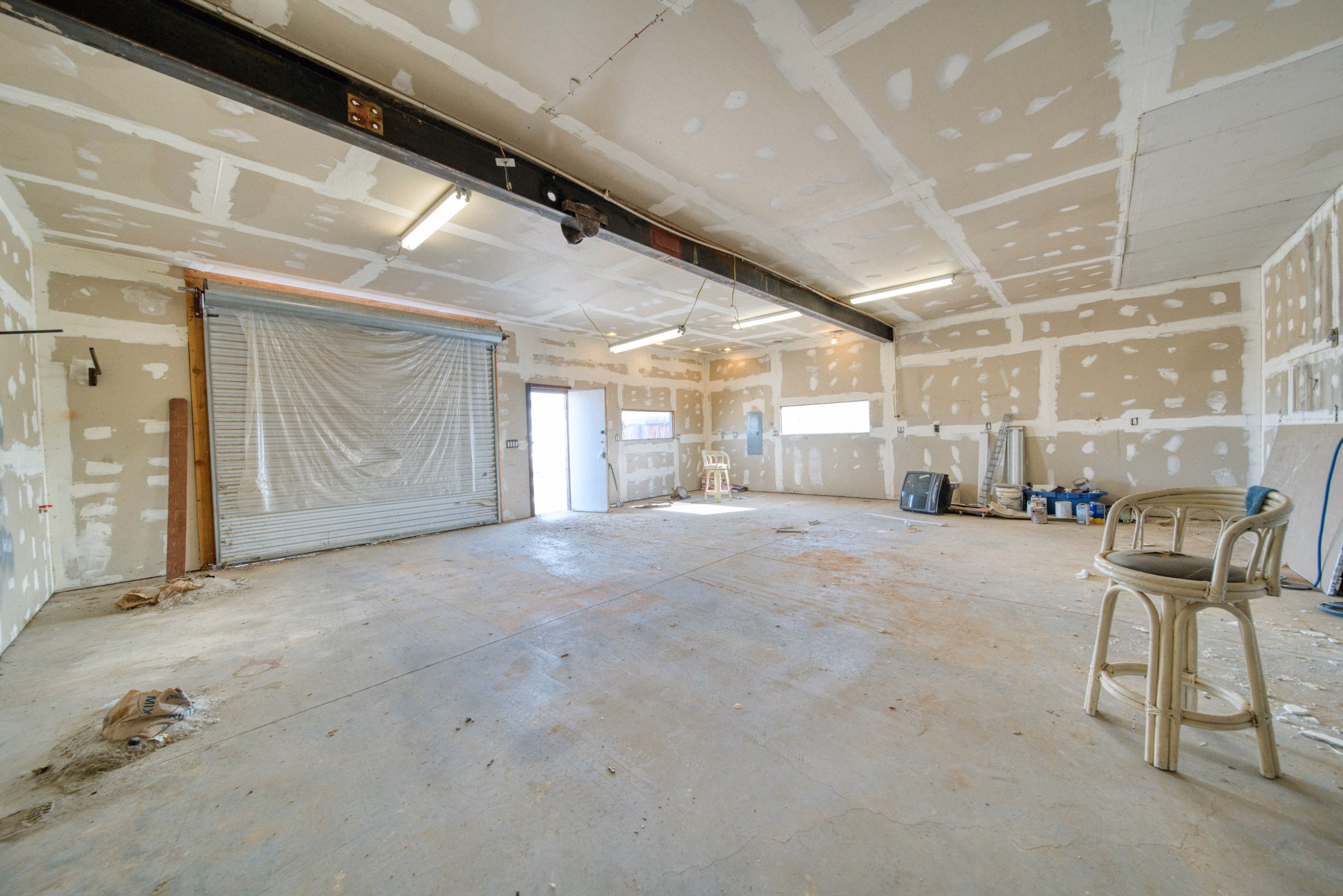Empty warehouse space with unfinished drywall, a concrete floor, two chairs, and a partially covered garage door.