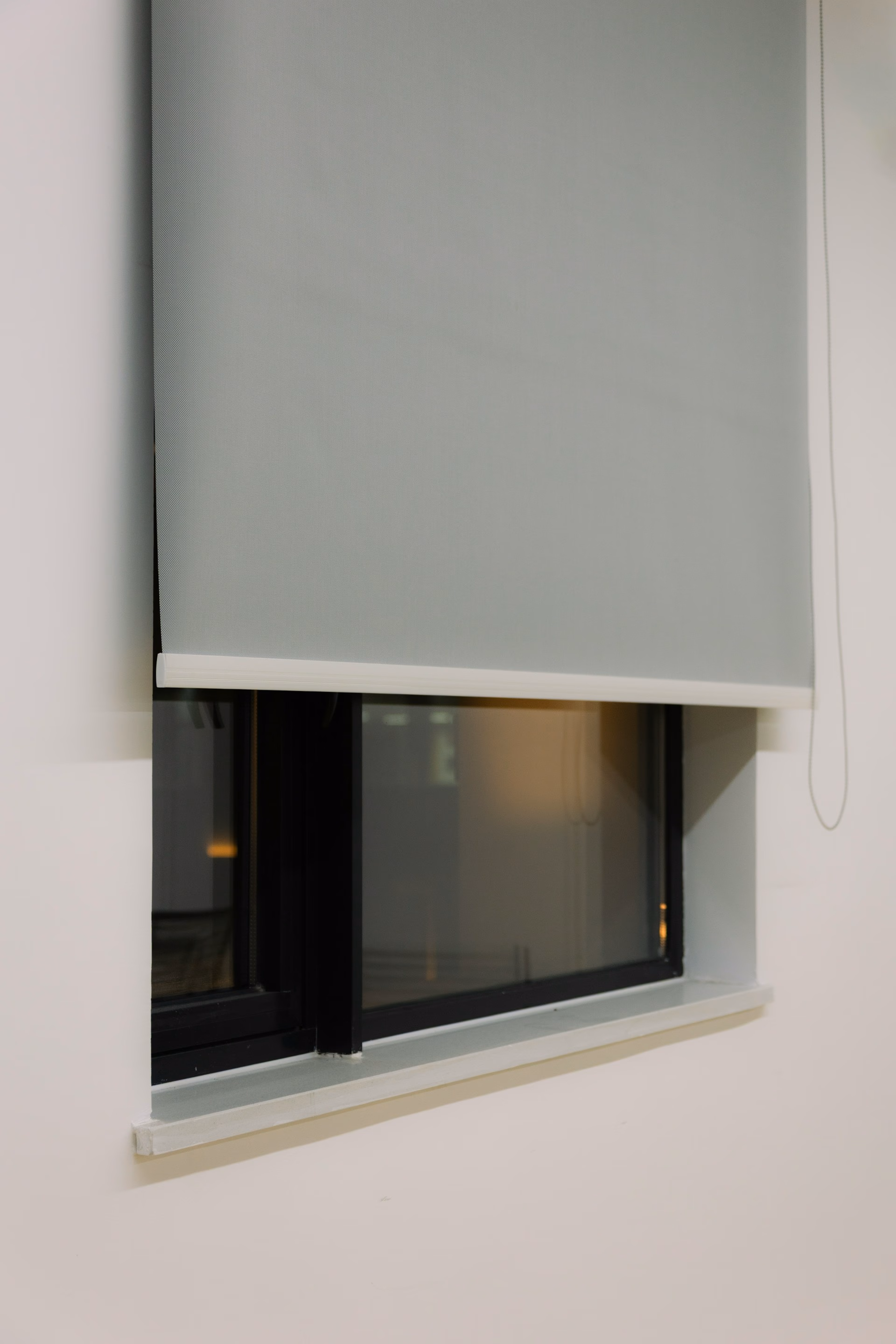 A partially lowered light gray roller blind over a black-framed window inside a beige wall.