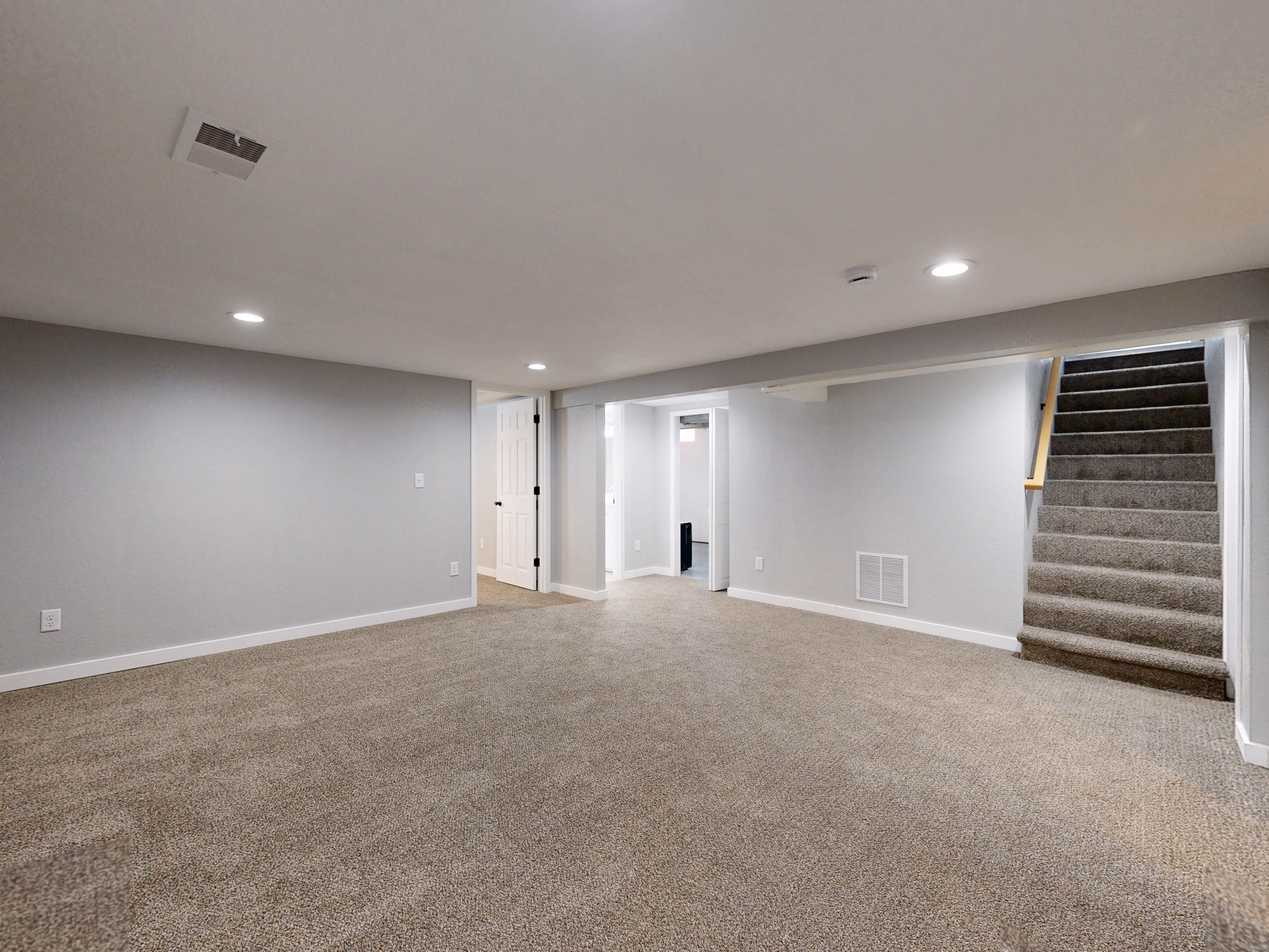 Empty basement room with beige carpet, gray walls, recessed ceiling lights, staircase, and open doors.